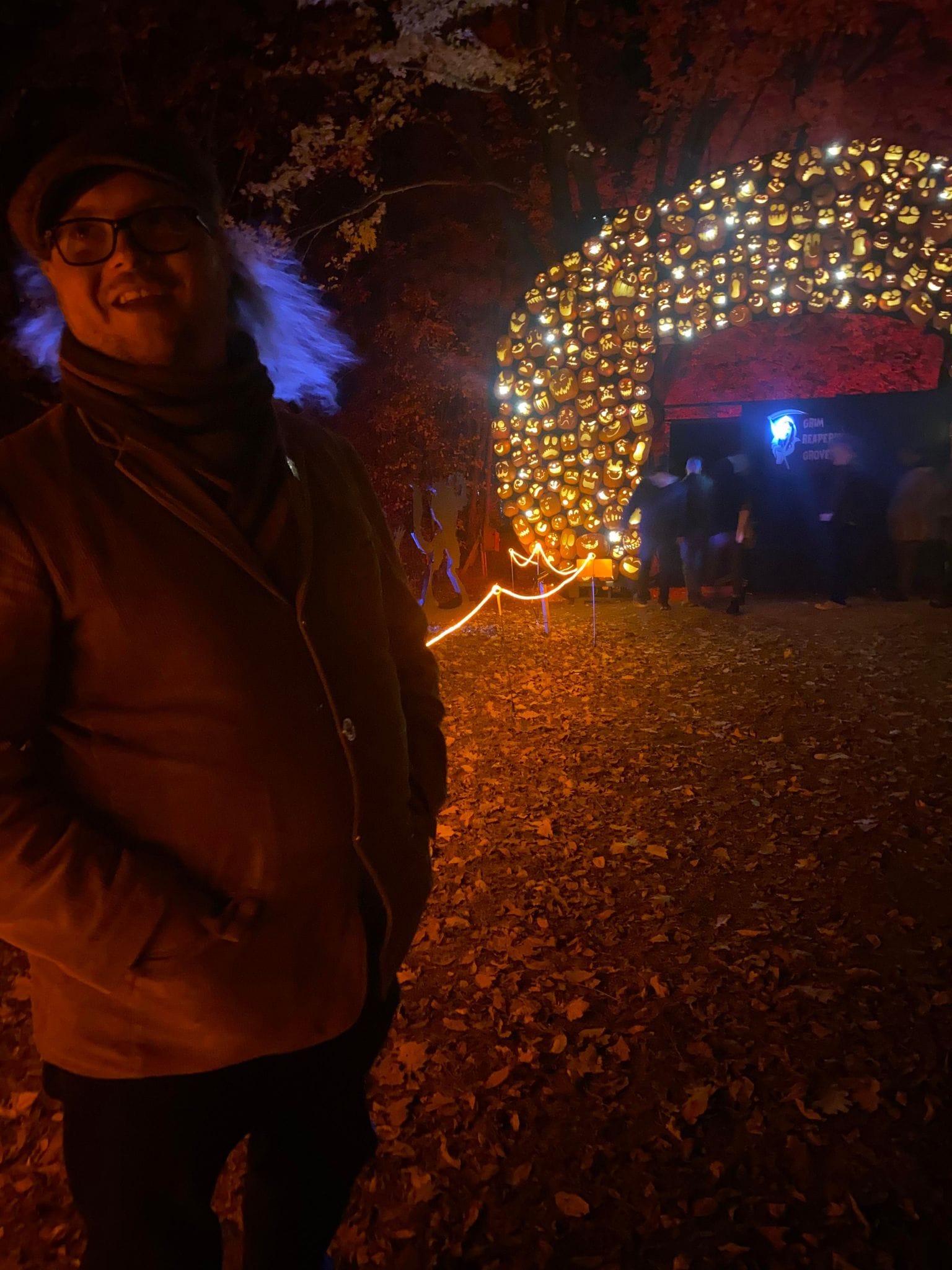 A man stands in front of an arch made of jack-o-lanturns, his hair looks purple in the light, Photo 1